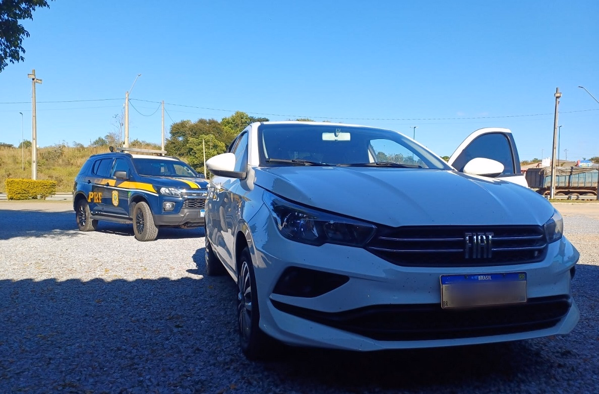 Fiat Cronos branco em rodovia do Sul de Minas após a recuperação de veículo furtado. Ao fundo, viatura da PRF e caminhões na Fernão Dias.