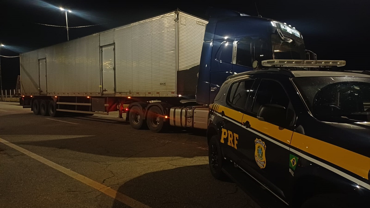 Imagem mostra agentes da PRF ao lado de carreta apreendida na BR-381 em Lavras, contendo mercadorias estrangeiras sem nota fiscal