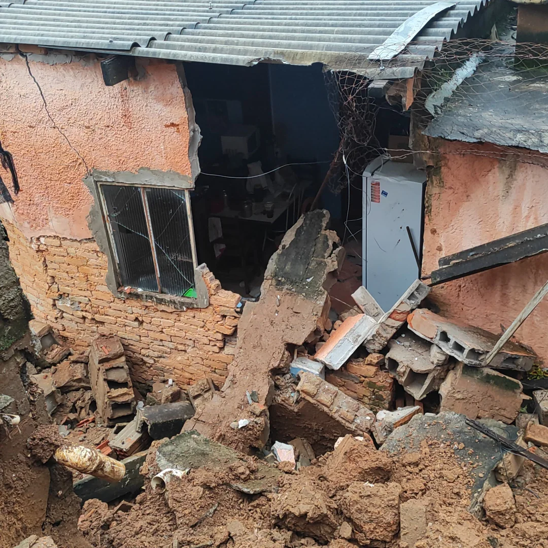 Fachada de residência atingida após muro desabar em Lavras, mostrando escombros e interdição da Defesa Civil.