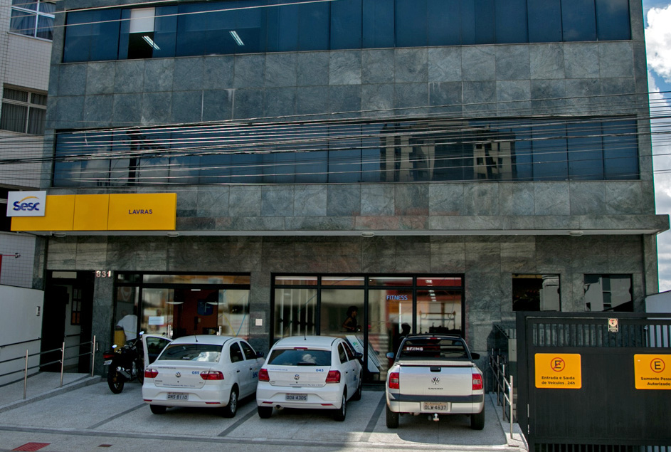 Fotografia colorida da fachada principal da unidade do Sesc em Lavras, Minas Gerais. A imagem mostra a entrada do prédio com o logo oficial do Sesc e a calçada acessível